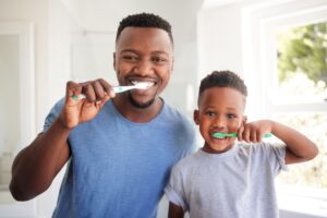 Man in blue shirt brushing his teeth in the bathroom with a boy in a gray shirt