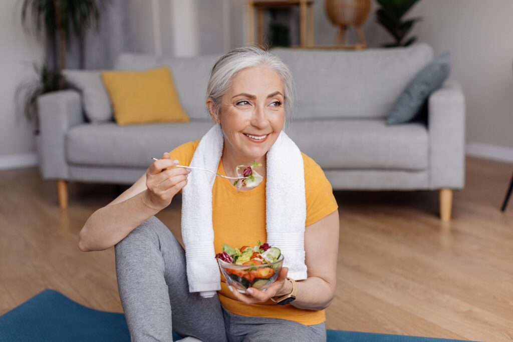 Woman with white hair in workout clothes eating salad on yoga mat